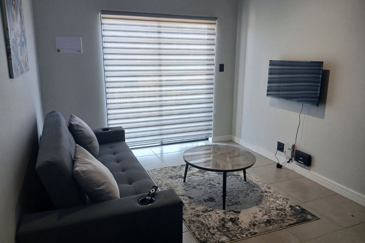 A compact living room featuring tiled flooring, neutral wall finishes, and a large window fitted with modern roller blinds.