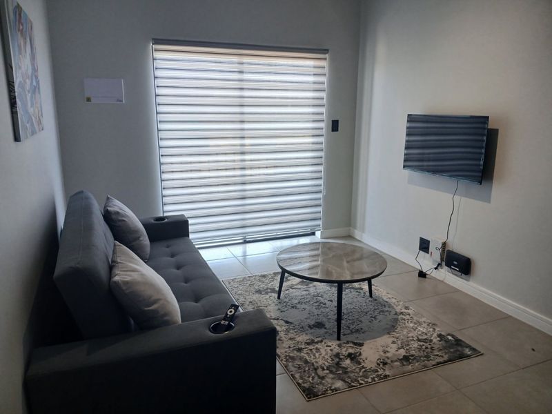A compact living room featuring tiled flooring, neutral wall finishes, and a large window fitted with modern roller blinds.