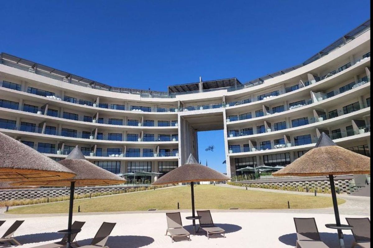 A modern, curved multi-story residential building featuring private balconies with glass balustrades, overlooking a landscaped lawn and sandy recreational area.