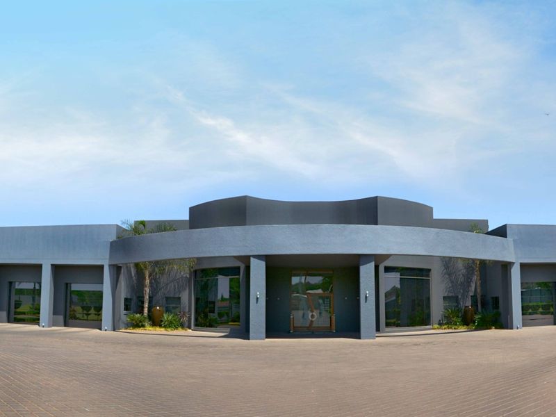 A modern, architecturally striking front facade featuring a prominent curved entrance portico, extensive glass windows, and a spacious paved driveway.