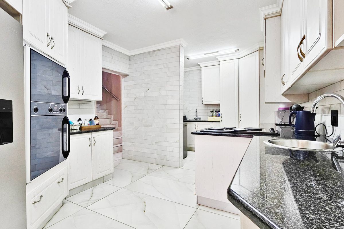 A bright, modern kitchen featuring white cabinetry, granite countertops, tiled backsplash, and large format floor tiles with a view toward the staircase.
