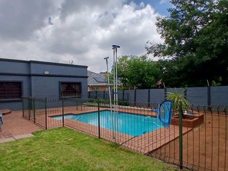 A backyard pool area featuring a paved deck, perimeter safety fencing, and a well-maintained lawn, set against a private boundary wall.