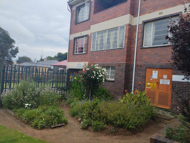 Exterior view of a brick apartment building featuring steel security windows, perimeter fencing, and an integrated electrical substation enclosure at ground level.