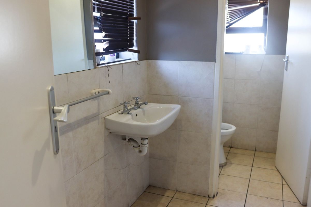 A functional commercial bathroom featuring a wall-mounted basin, tiled surfaces, and a separate toilet area, requiring minor maintenance and cosmetic updates.