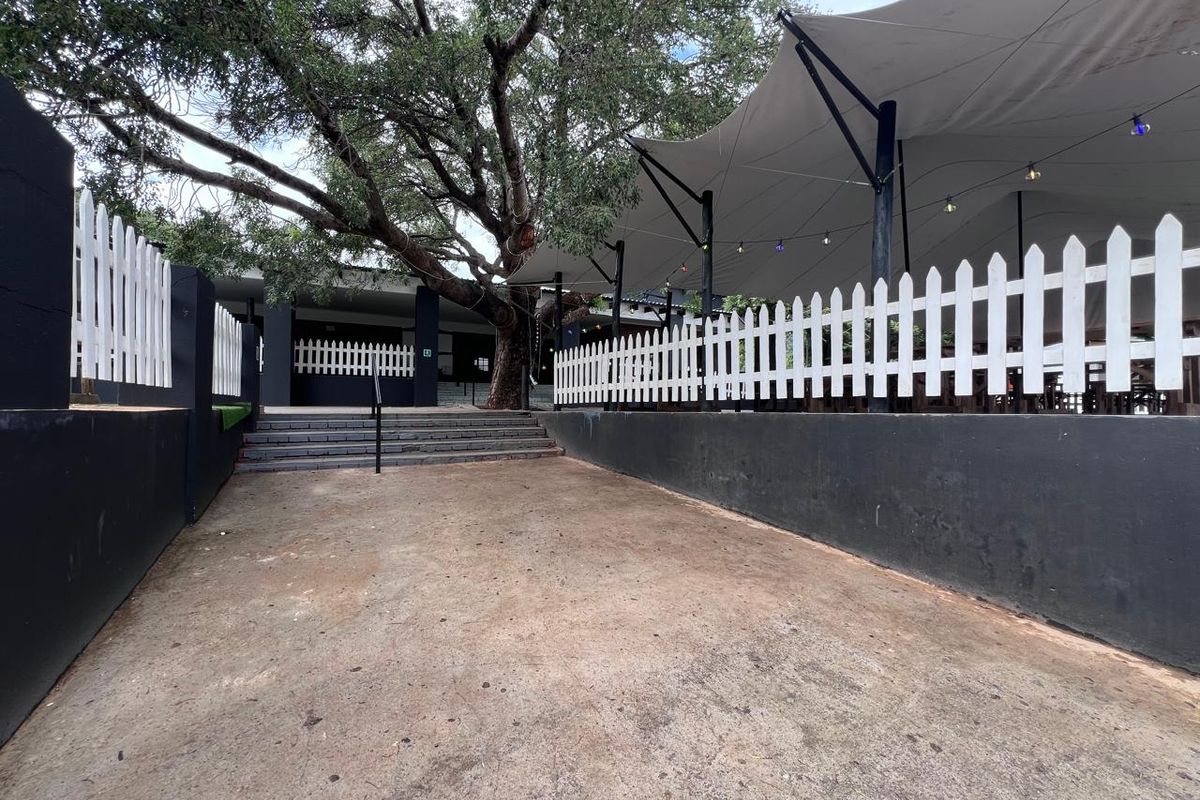 Outdoor commercial patio area featuring a tensioned fabric canopy, white picket fencing, and concrete stairs leading to the main building entrance.