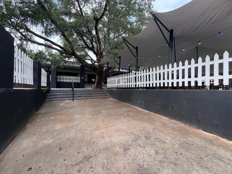 Outdoor commercial patio area featuring a tensioned fabric canopy, white picket fencing, and concrete stairs leading to the main building entrance.
