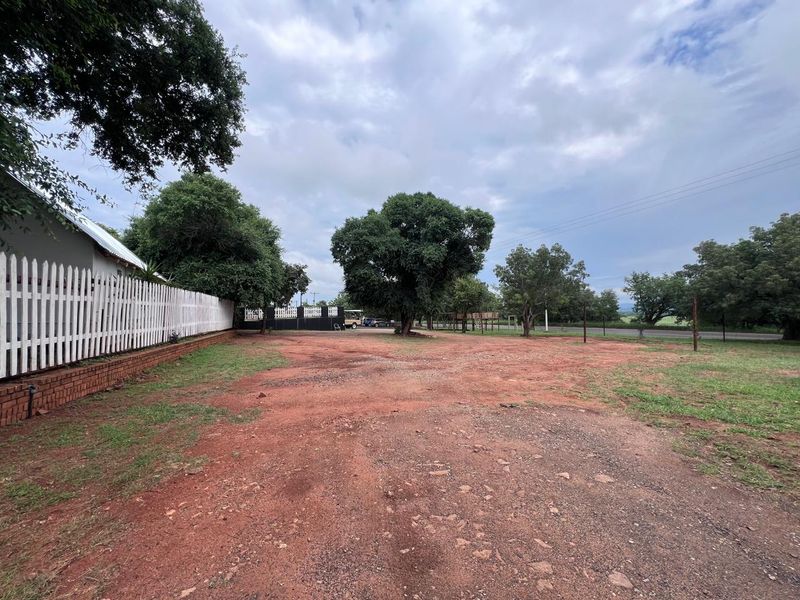 A wide view of the property frontage featuring a gravel driveway, mature trees, and a white picket fence along the boundary.