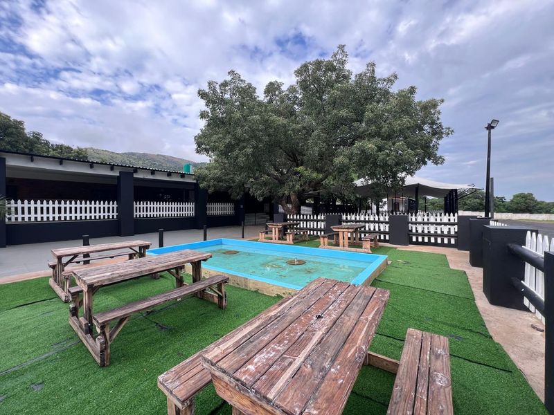 Outdoor commercial entertainment area featuring a central fountain, artificial turf surfacing, and perimeter picket fencing with access to a covered patio space.