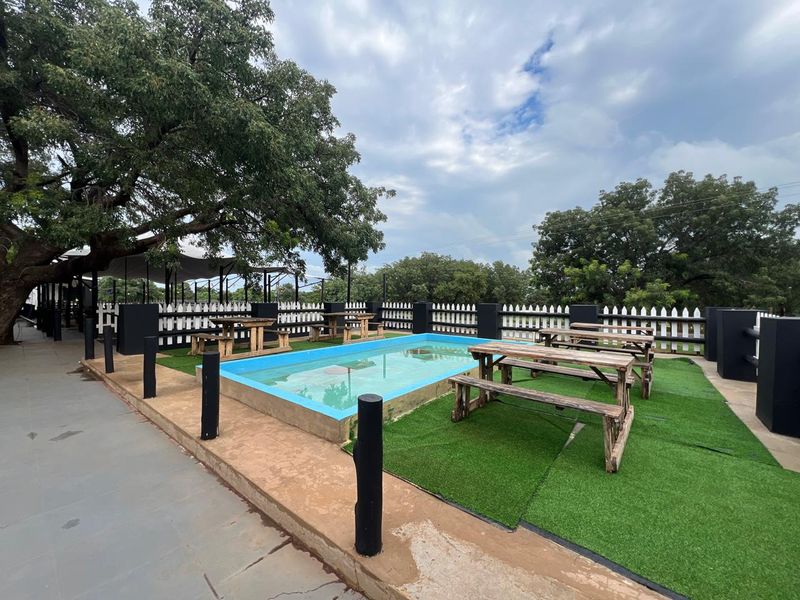 Outdoor commercial pool area featuring a small swimming pool, artificial turf surfacing, and perimeter picket fencing under a large shade tree.