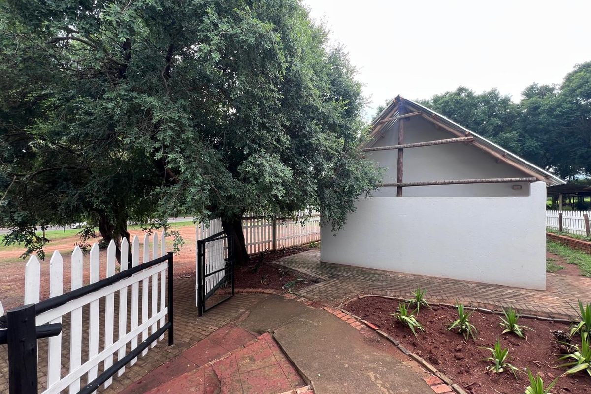 A rear exterior view showing a paved walkway, garden beds, and a white picket fence leading to an unfinished building structure.
