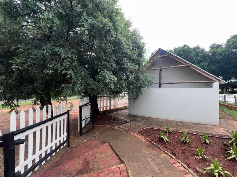 A rear exterior view showing a paved walkway, garden beds, and a white picket fence leading to an unfinished building structure.