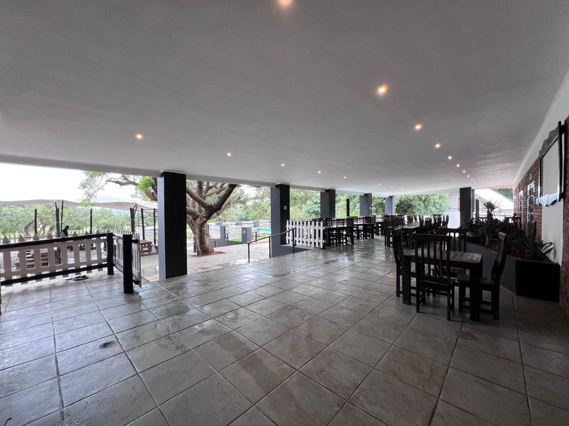 A spacious covered outdoor patio area featuring tiled flooring, recessed lighting, and structural columns, overlooking a garden and pool area.