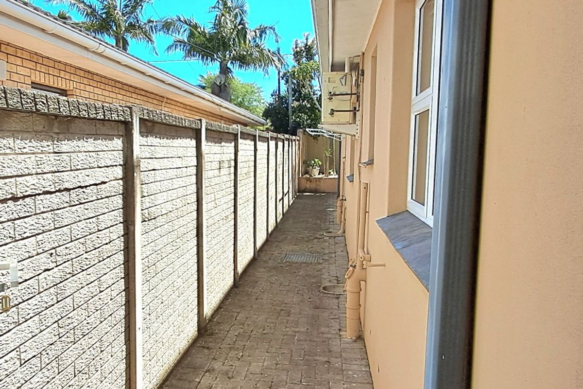 A narrow side passage featuring a paved walkway, precast concrete boundary wall, and external building services including plumbing and air conditioning units.