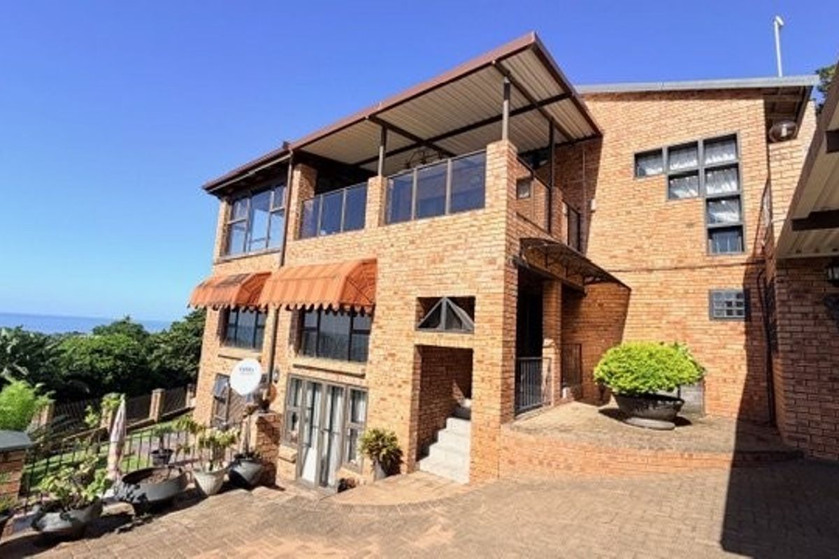 A multi-level face brick residence featuring a covered balcony with glass balustrades, paved driveway, and scenic coastal views.