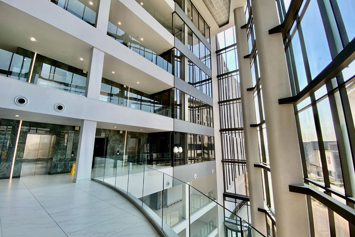 A modern, multi-level commercial atrium featuring expansive glass curtain walls, marble-clad elevator lobbies, and sleek glass balustrades in a high-quality, contemporary office building.