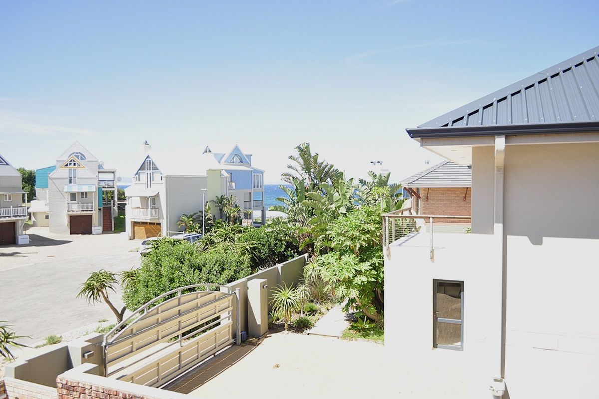 An elevated view of the rear exterior, featuring a paved driveway, boundary wall, metal gate, and a balcony overlooking the surrounding coastal neighborhood.