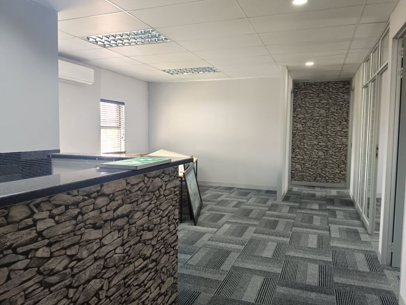 A modern commercial office space featuring a stone-clad reception desk, carpet tiles, suspended ceilings with recessed lighting, and glass partition walls.