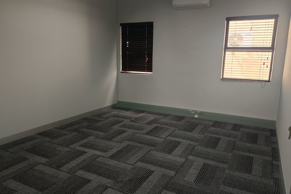 A clean, empty office space featuring grey carpet tiles, split-system air conditioning, and windows fitted with venetian blinds.