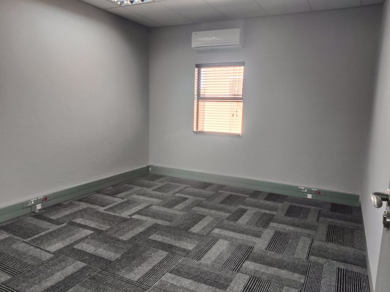 A clean, empty office space featuring grey carpet tiles, a suspended ceiling, wall-mounted air conditioning, and integrated electrical trunking along the baseboards.