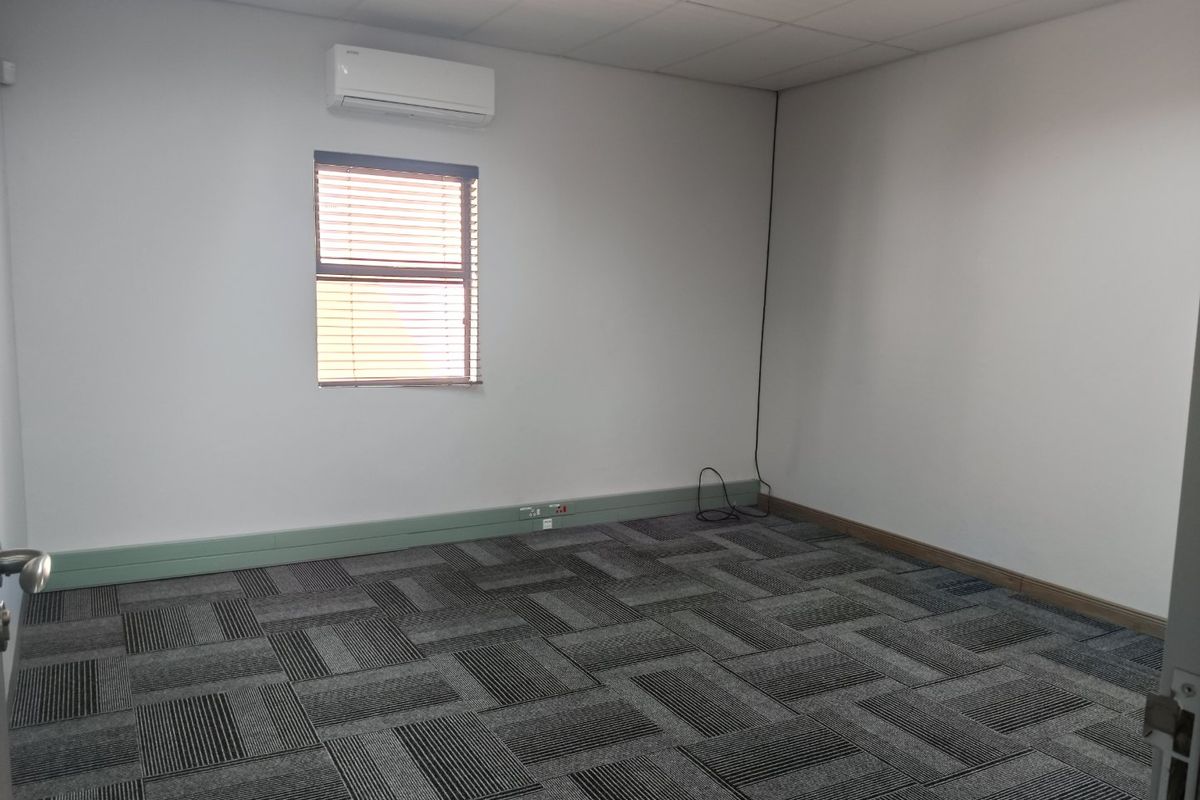 A clean, vacant office space featuring carpet tiles, a suspended ceiling, and a wall-mounted air conditioning unit.