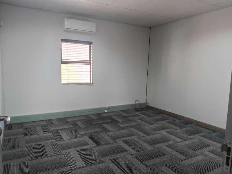 A clean, vacant office space featuring carpet tiles, a suspended ceiling, and a wall-mounted air conditioning unit.