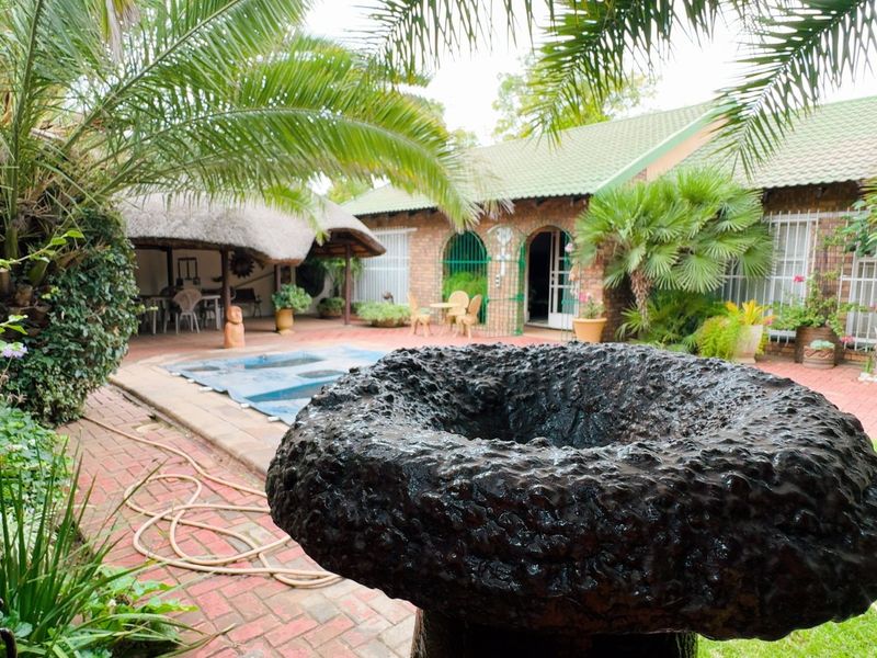 Outdoor pool area featuring a swimming pool, thatched lapa, and established garden with brick paving and a stone water feature.