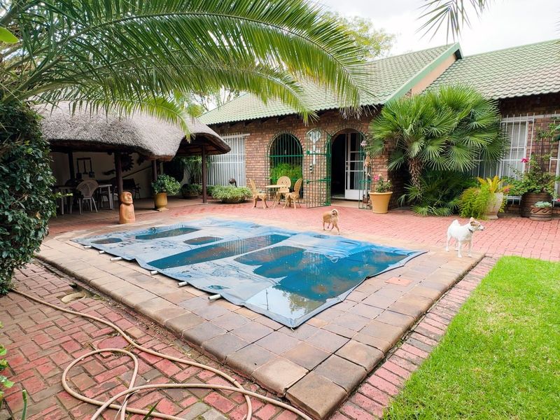 Outdoor pool area featuring a swimming pool with cover, brick paved patio, and a thatched lapa, set against a brick house facade.