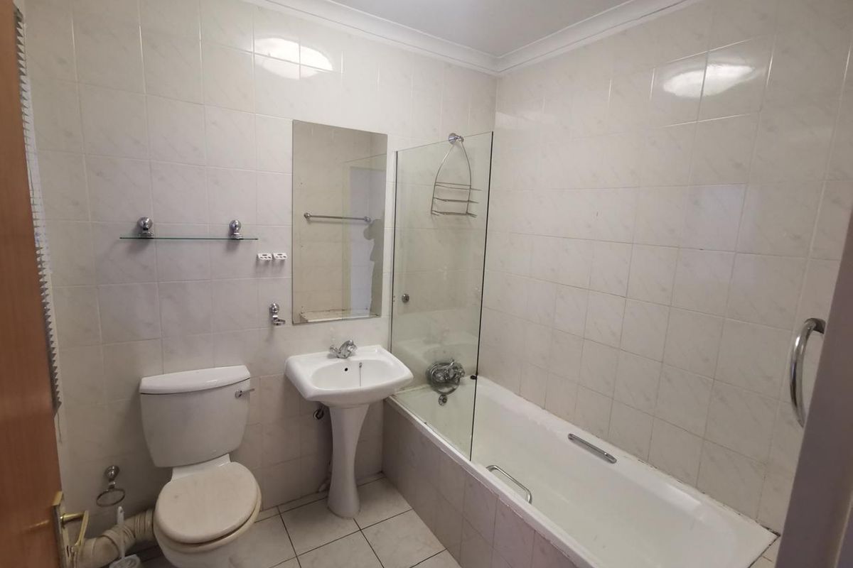 A functional bathroom featuring a pedestal basin, toilet, and a bathtub with a glass shower screen, finished with neutral wall and floor tiling.
