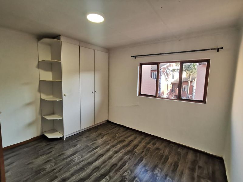 A bright, empty bedroom featuring modern laminate flooring, a built-in wardrobe with open shelving, and a window overlooking the complex.