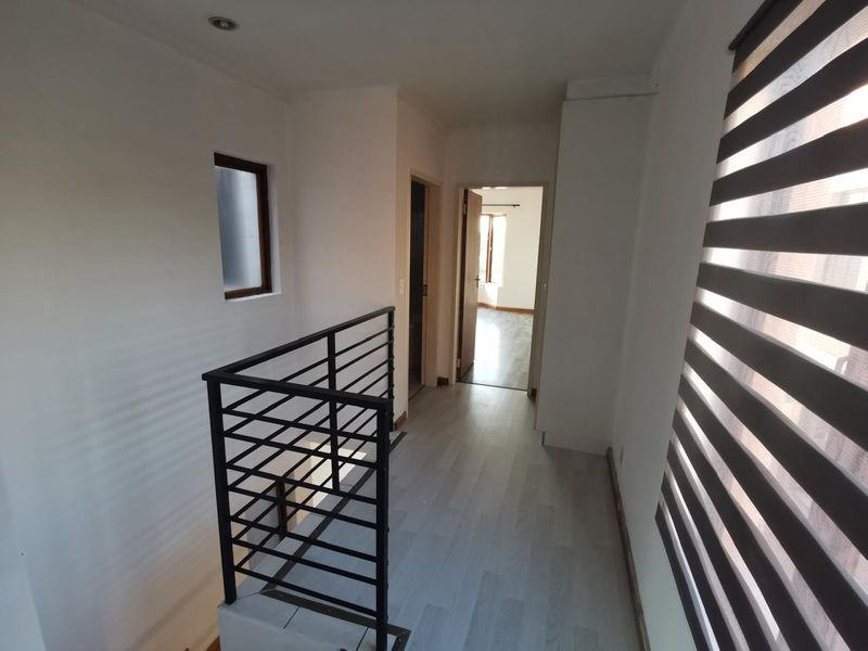 A bright, empty upper-level hallway featuring modern laminate flooring, a black metal balustrade, and clean white walls with recessed lighting.
