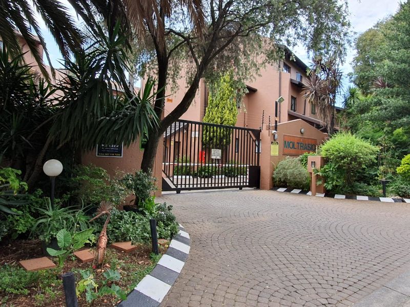 The entrance to Moltrasio apartment complex featuring a secure gated access point, paved driveway, and established perimeter landscaping.