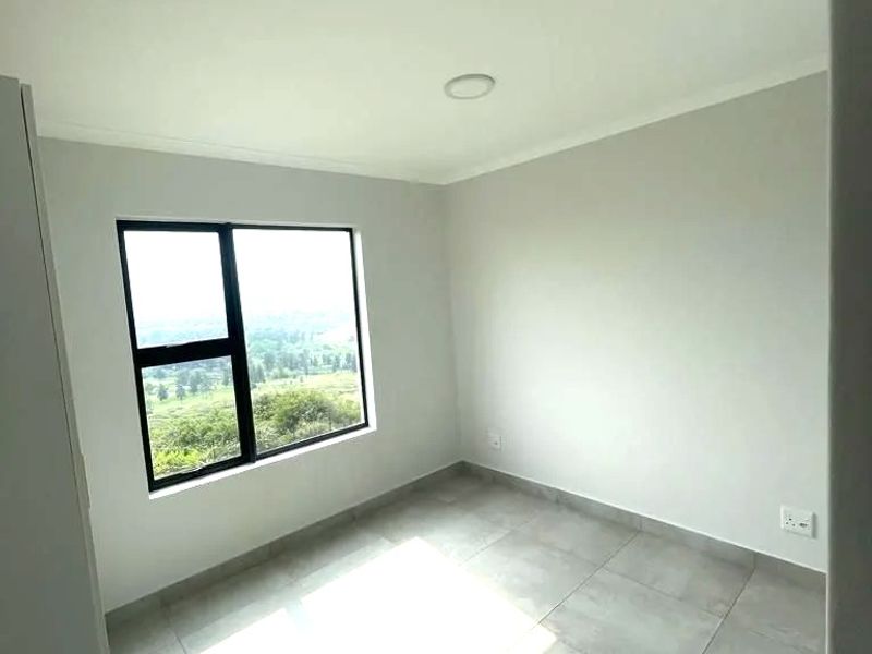 A bright, empty bedroom featuring neutral tiled flooring, clean walls, and a large window with natural light and scenic views.