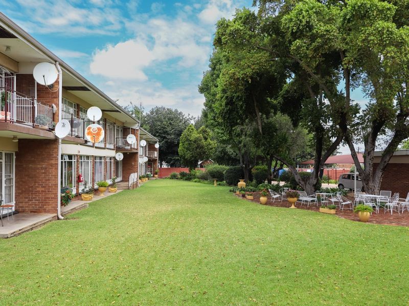 A view of the rear apartment building facade featuring brick construction, private balconies, and a well-maintained communal garden area with mature trees.
