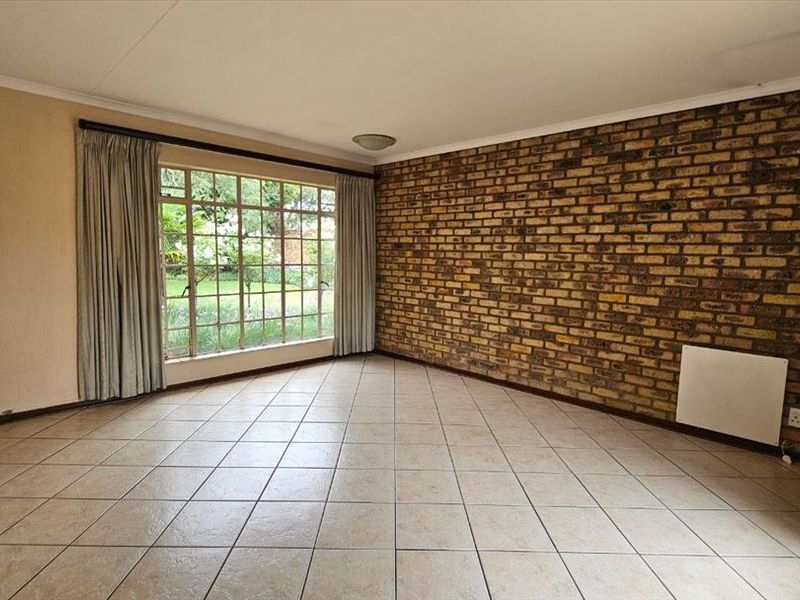 A spacious, empty living room featuring a prominent exposed brick wall, neutral tiled flooring, and a large window overlooking the garden.