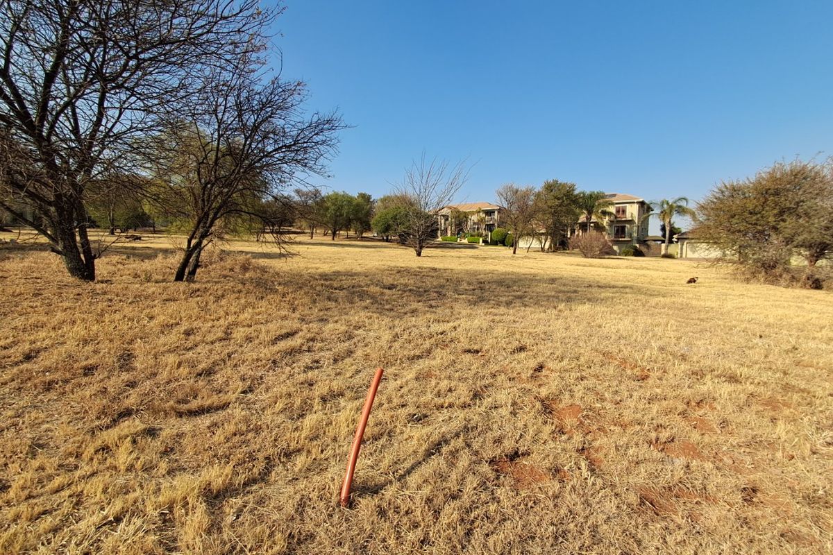 A wide-angle view of a vacant residential plot featuring dry grass, mature trees, and a survey marker in the foreground.