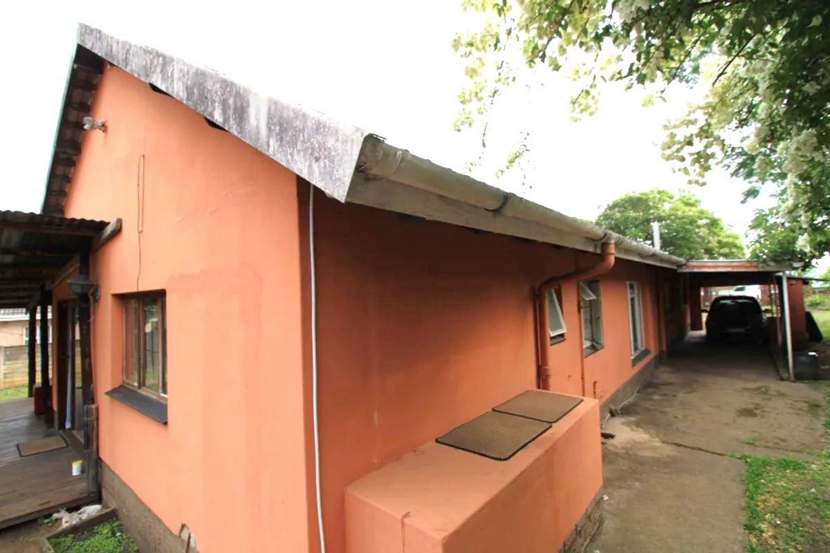 Rear exterior view of the house showing a concrete driveway, carport, and timber decking area with weathered wall finishes and roof fascia.