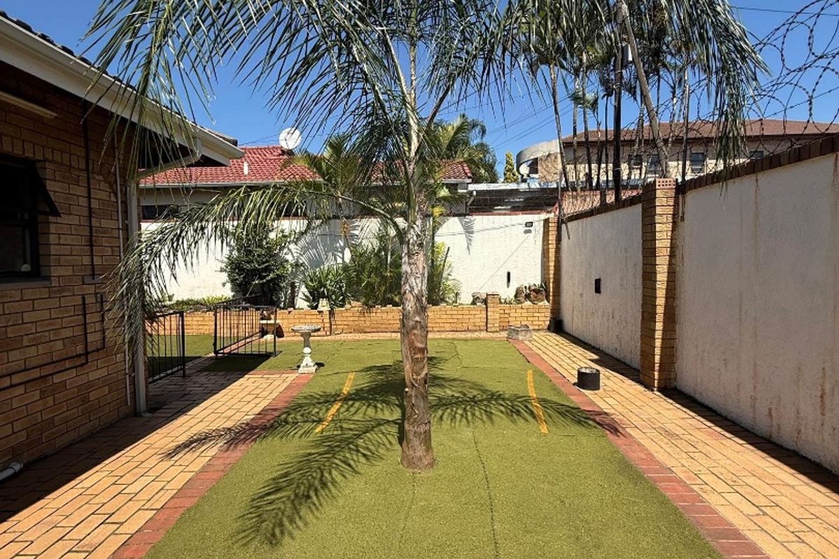 A rear exterior area featuring a paved walkway, artificial turf section, and a central palm tree enclosed by a brick boundary wall with security fencing.