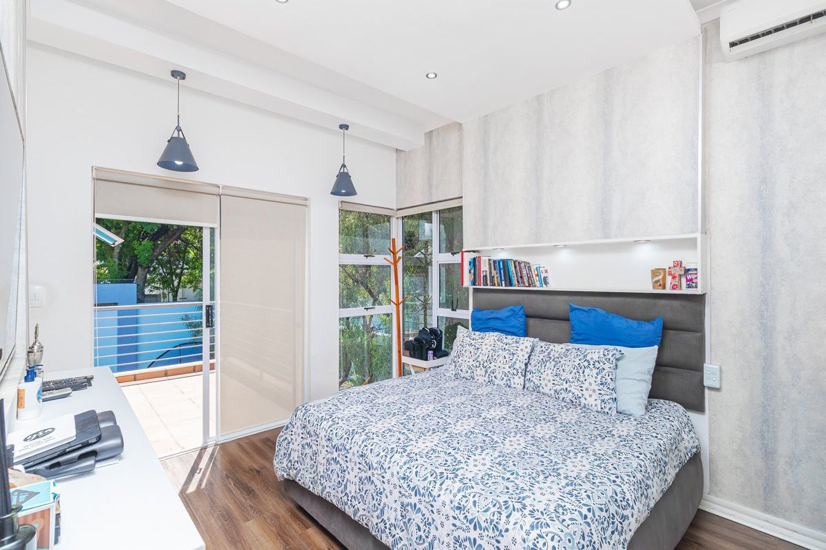 A bright bedroom featuring wood-look flooring, built-in headboard with integrated shelving, recessed lighting, air conditioning unit, and sliding glass doors leading to a private balcony.