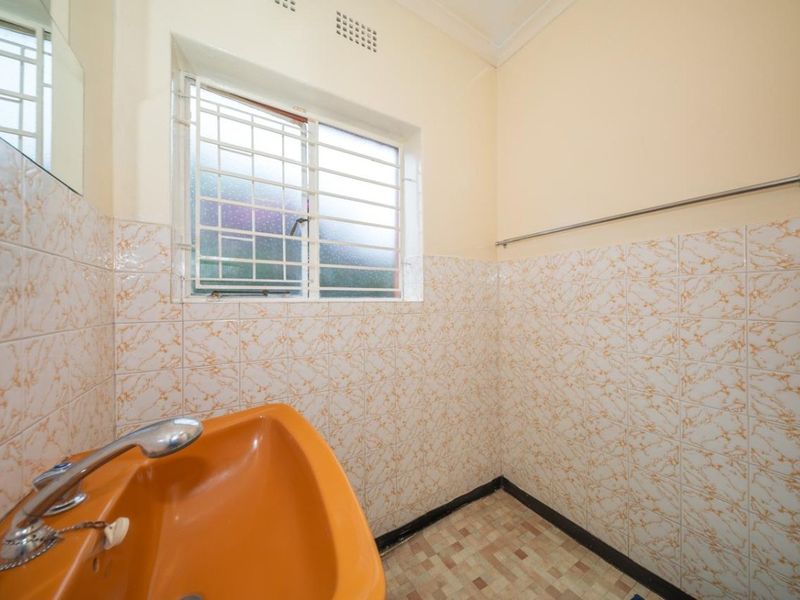 A compact bathroom featuring a vintage orange pedestal basin, patterned wall tiles, and security bars on the window.