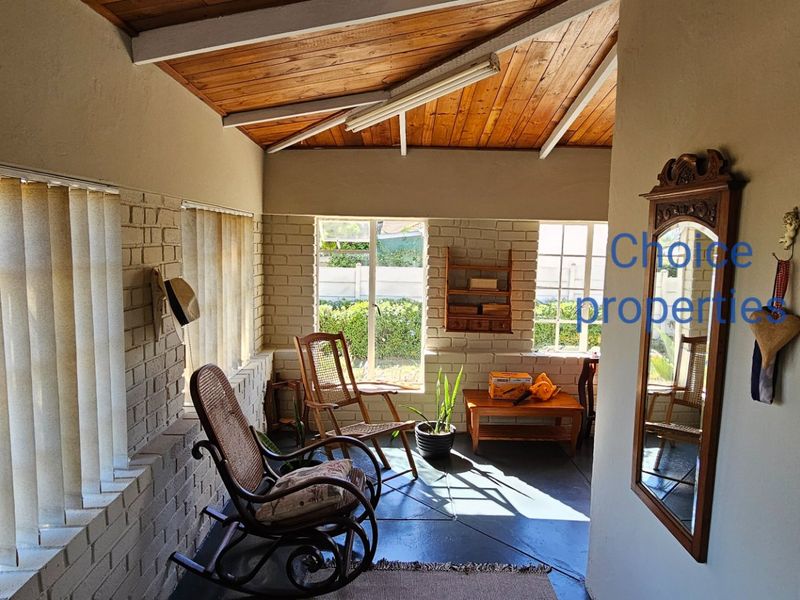 A sunlit enclosed patio featuring exposed timber ceilings, painted brick walls, and large steel-framed windows overlooking the garden.
