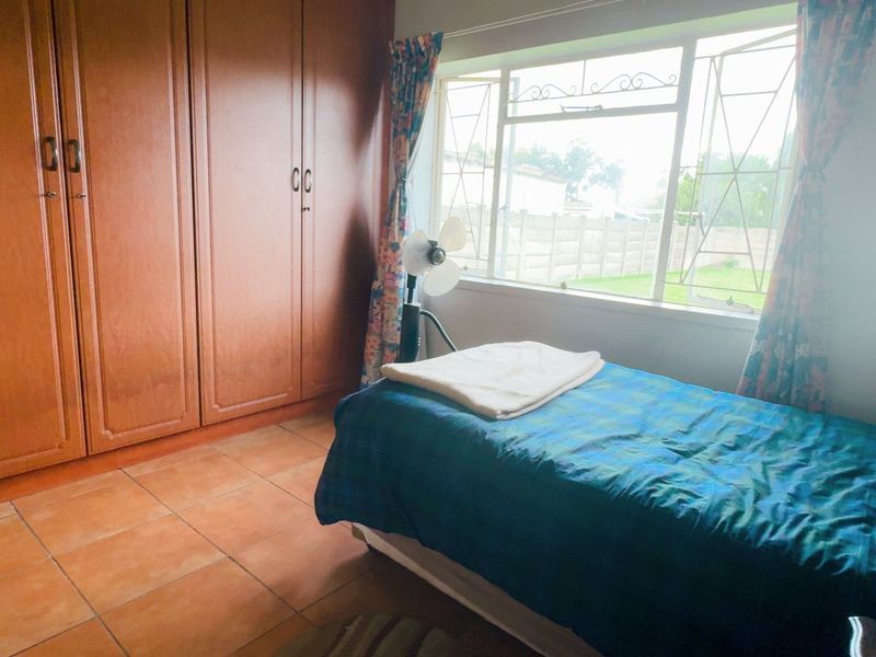 A functional bedroom featuring built-in wooden cupboards, tiled flooring, and a window with security bars overlooking the rear garden.