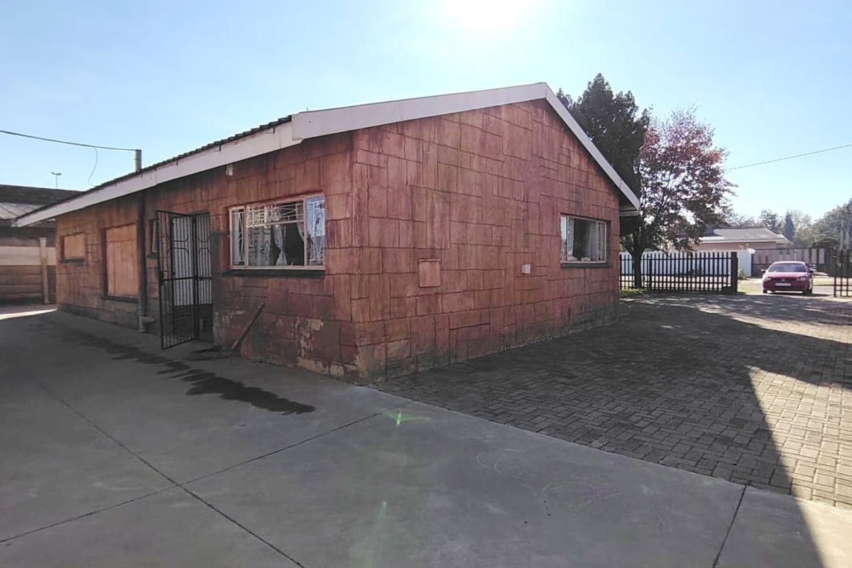 Exterior view of a detached house featuring a paved driveway, security gate, and weathered masonry walls with some windows boarded up.