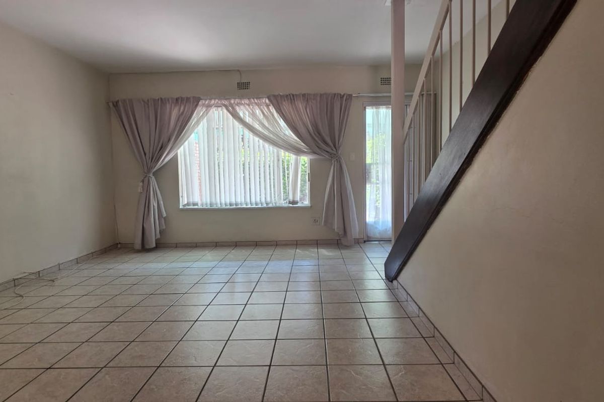 An empty, open-plan living area featuring tiled flooring, a staircase, and natural light from a large window and glass door.