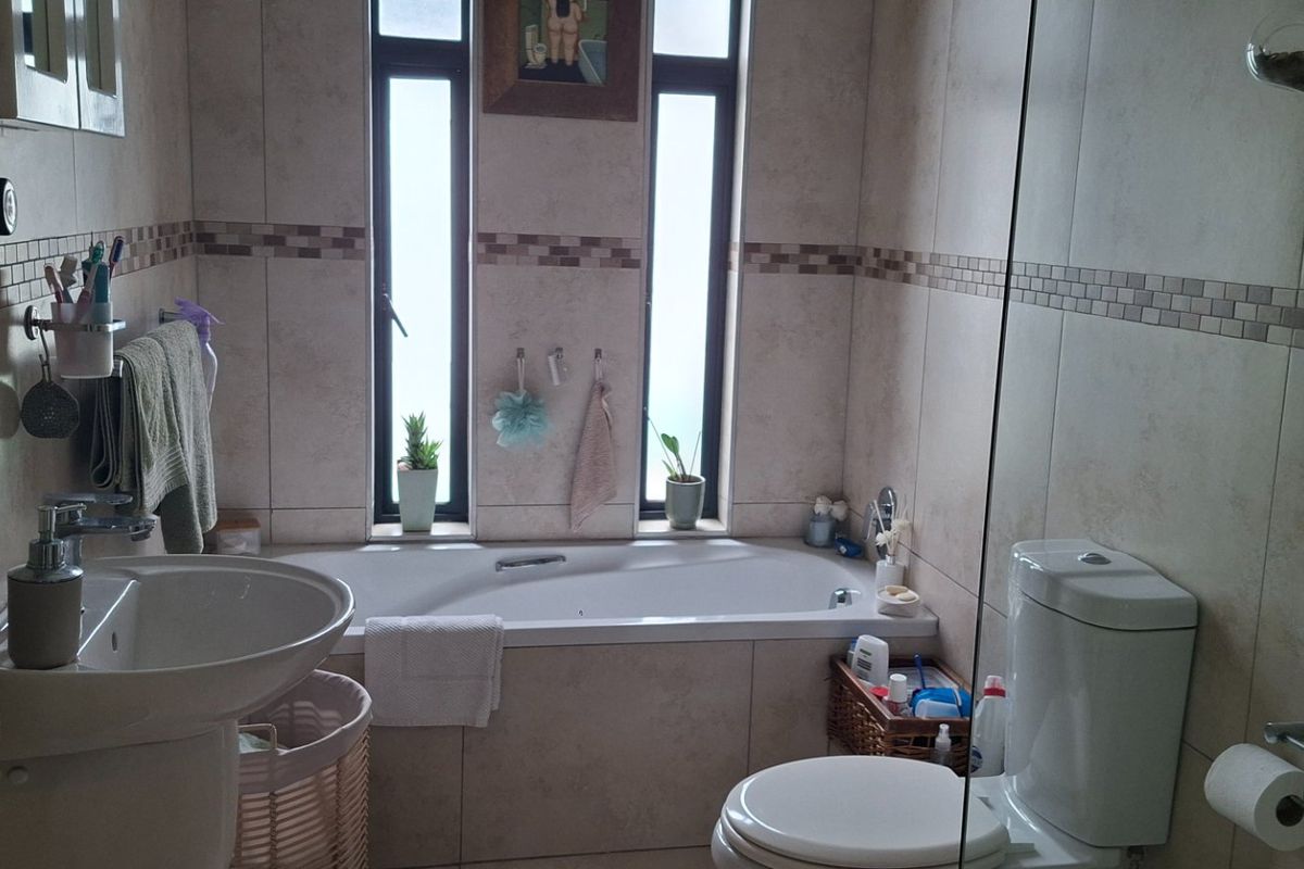 A functional bathroom featuring a built-in bathtub, pedestal basin, and dual-flush toilet with neutral wall and floor tiling.