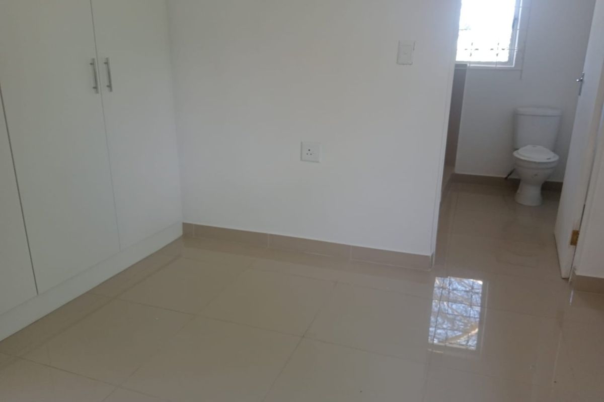 A clean, empty bedroom featuring built-in cupboards, neutral tiled flooring, and an adjoining en-suite toilet area.