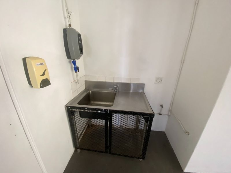 A functional utility area featuring a stainless steel sink with a metal mesh cabinet base, wall-mounted dispensers, and exposed industrial-style piping.