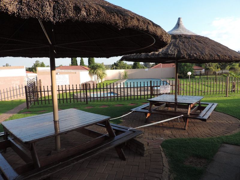 Communal outdoor pool area featuring a paved patio, thatched umbrellas, and perimeter fencing within a landscaped garden setting.