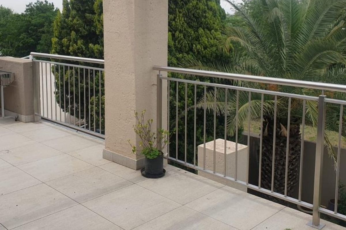 A spacious balcony featuring modern stainless steel balustrades, neutral tiled flooring, and a sturdy concrete support pillar overlooking the garden area.