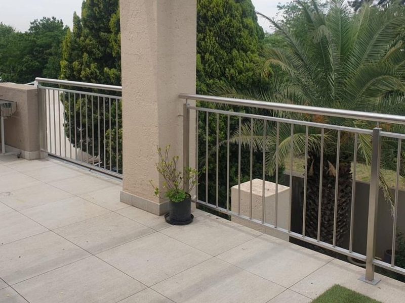 A spacious balcony featuring modern stainless steel balustrades, neutral tiled flooring, and a sturdy concrete support pillar overlooking the garden area.