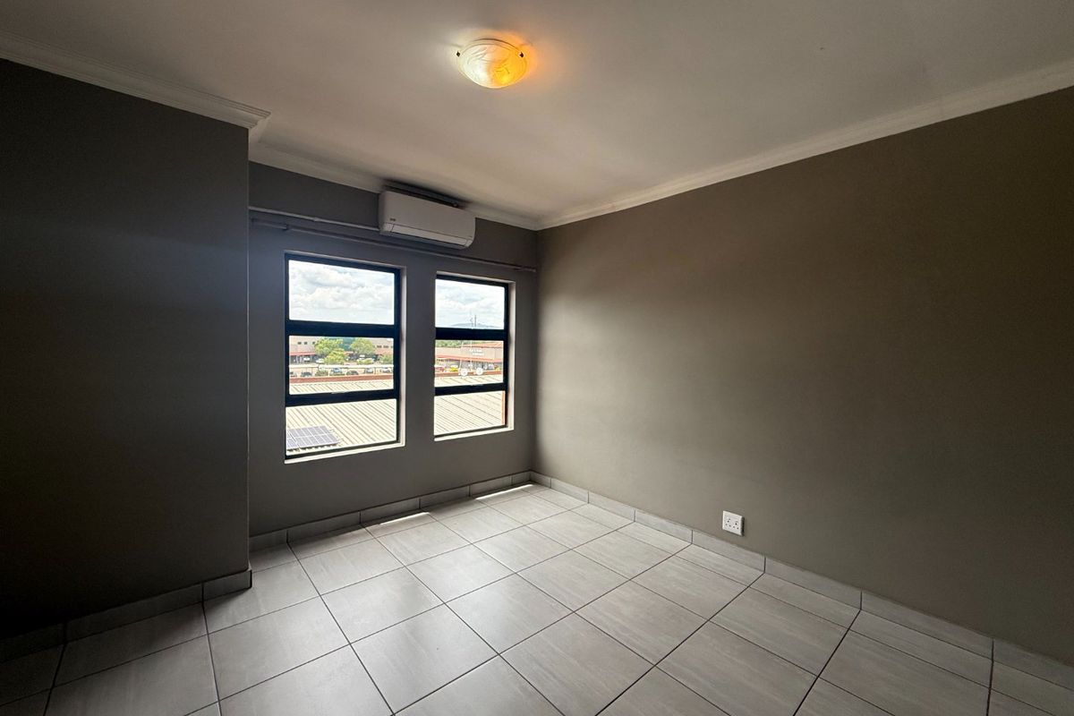 A clean, empty bedroom featuring neutral tiled flooring, an installed air conditioning unit, and large windows providing natural light.